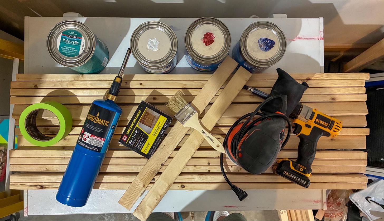 Flat-lay of the tools and materials to build a DIY wood American flag — raw hardwood slats, wood stain, spray paint, a star stencil, torch, clamps and a drill